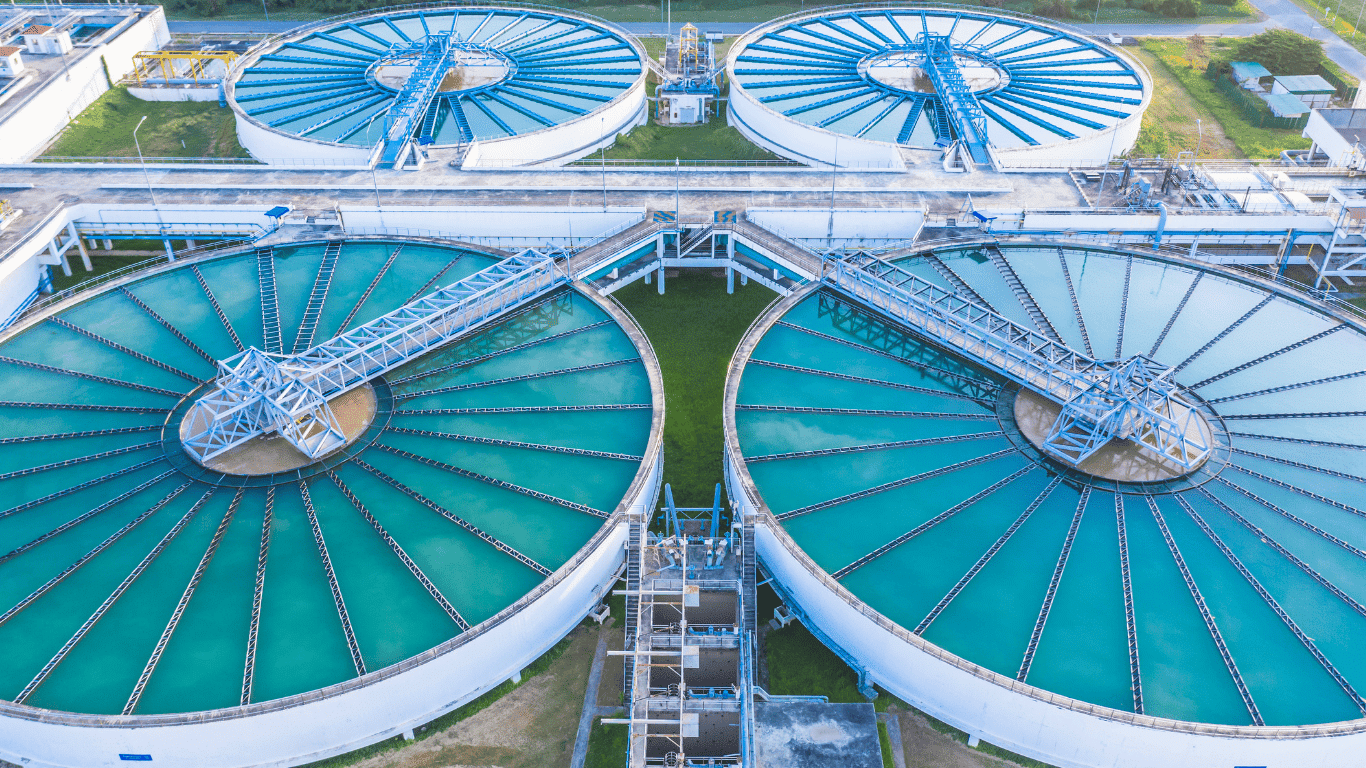 Aerial view of a modern water treatment plant showing multiple circular basins at a large treatment plant where water treatment transforms raw water into clean drinking water for the public water supply. The first step of the drinking water process begins as raw water is pumped through pipes into the plant, illustrated like an arrow slideshow explaining the full system from intake to tap. In the coagulation flocculation stage, chemicals such as ferric sulfate are added, creating positive charges that attract negatively charged particles, forming floc particles from microscopic particles, sediment, and suspended solids. These larger particles combine and settle during the sedimentation process, helping remove suspended solids from the water. The clarified clear water flows onward while heavy sediment sinks to the bottom. Filtration layers of sand and gravel further remove remaining particles before backwash water is used to clean the filters. Advanced treatment includes granular activated carbon to absorb contaminants, followed by ozone disinfection using ozone to destroy harmful bacteria and viruses. Chlorine disinfection and secondary disinfection with chlorine ensure long-lasting protection as water moves through distribution pipes. This complete water treatment process delivers safe drinking water from source to tap, ensuring public health through effective removal of particles, pathogens, and contaminants.